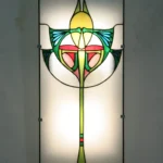 Détail d'une applique murale ou d'un panneau en vitrail de style Art Nouveau. La pièce présente un motif floral stylisé et symétrique sur fond dépoli et rétroéclairé. Les couleurs dominantes sont le jaune, le vert vif, le rouge, le rose saumon, et le bleu turquoise, encadrées par des lignes de plomb noires. Des textures de verre striées sont visibles sur les bords inférieurs du motif, évoquant des ailes ou des feuilles