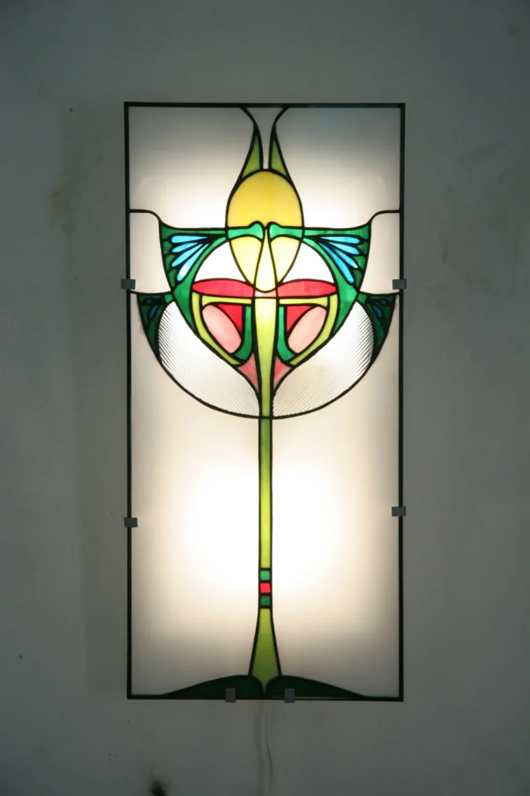 Détail d'une applique murale ou d'un panneau en vitrail de style Art Nouveau. La pièce présente un motif floral stylisé et symétrique sur fond dépoli et rétroéclairé. Les couleurs dominantes sont le jaune, le vert vif, le rouge, le rose saumon, et le bleu turquoise, encadrées par des lignes de plomb noires. Des textures de verre striées sont visibles sur les bords inférieurs du motif, évoquant des ailes ou des feuilles