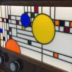 Coffre en bois foncé poli, de style Art Moderne, avec des formes arrondies sur les côtés. La façade principale est ornée d'un panneau de vitrail rétroéclairé. Le motif du vitrail est abstrait et géométrique, inspiré par le mouvement De Stijl (Piet Mondrian), composé de rectangles et de lignes noires sur fond blanc opalescent, intégrant des cercles colorés (jaune, bleu, orange, rouge). Le coffre possède deux tiroirs cachés sur les côtés et un couvercle supérieur qui s'ouvre pour révéler un miroir et un compartiment. Trois boutons ronds sont alignés sous le vitrail. L'éclairage LED interne diffuse une lumière douce à travers le vitrail.