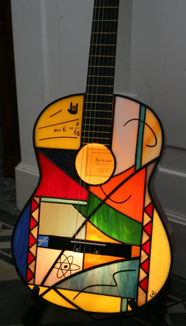 Lampe de sol artistique créée à partir d'une guitare acoustique transformée. La caisse de résonance de la guitare a été remplacée par un panneau en vitrail de style Tiffany, aux motifs abstraits et géométriques. Le vitrail intègre une palette de couleurs vives : bleu ciel, vert émeraude, rouge, orange, jaune, et verre irisé. Le corps en vitrail est éclairé par l'intérieur, illuminant le motif qui inclut de petits détails comme des notes de musique stylisées près de l'ouïe. La guitare est posée sur un support de sol noir sur un carrelage effet bois.