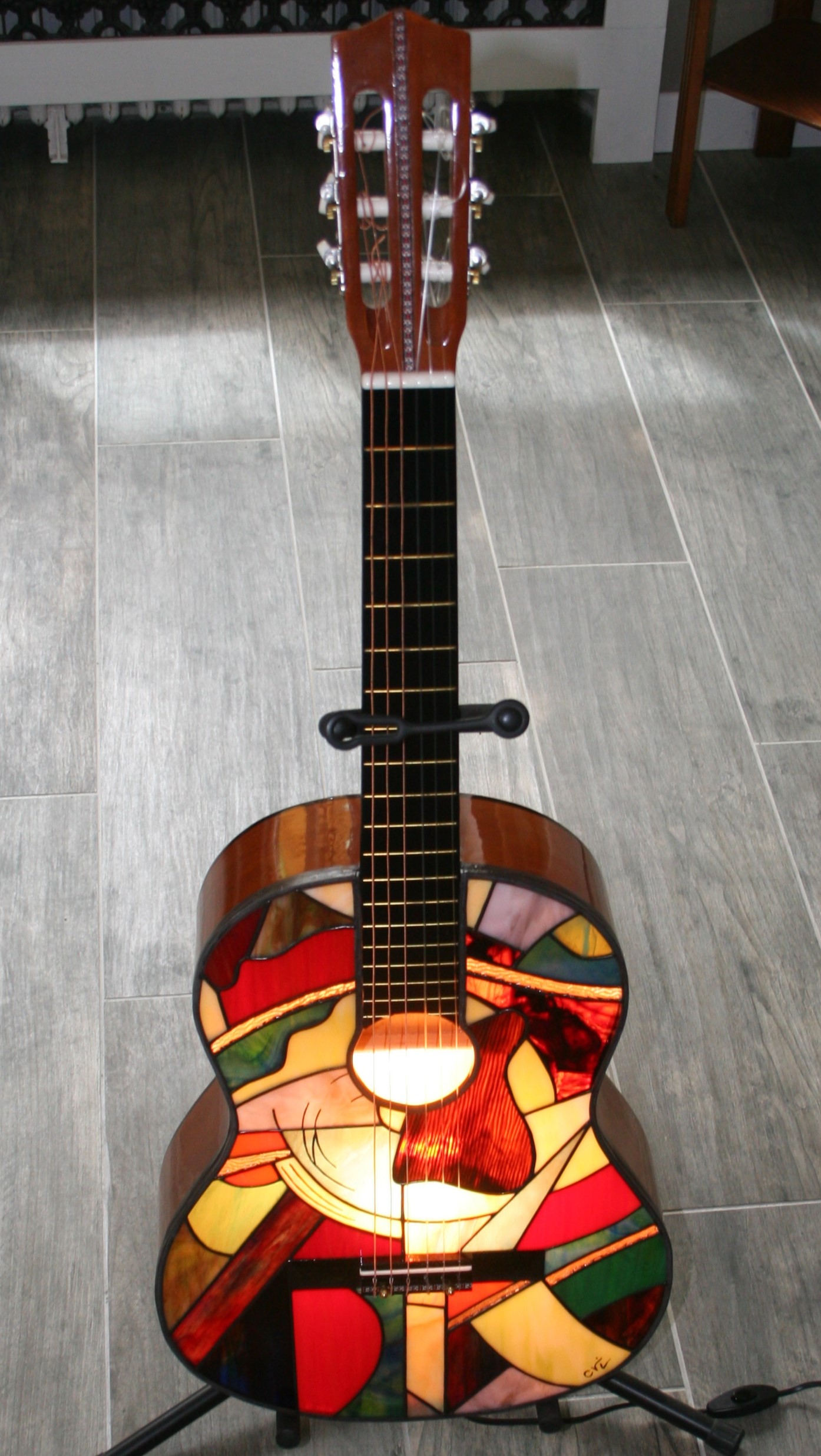 Lampe de sol artistique créée à partir d'une guitare acoustique transformée. La caisse de résonance de la guitare a été remplacée par un panneau en vitrail de style Tiffany, aux motifs abstraits et géométriques. Le vitrail intègre une palette de couleurs vives : bleu ciel, vert émeraude, rouge, orange, jaune, et verre irisé. Le corps en vitrail est éclairé par l'intérieur, illuminant le motif qui inclut de petits détails comme des notes de musique stylisées près de l'ouïe. La guitare est posée sur un support de sol noir sur un carrelage effet bois.