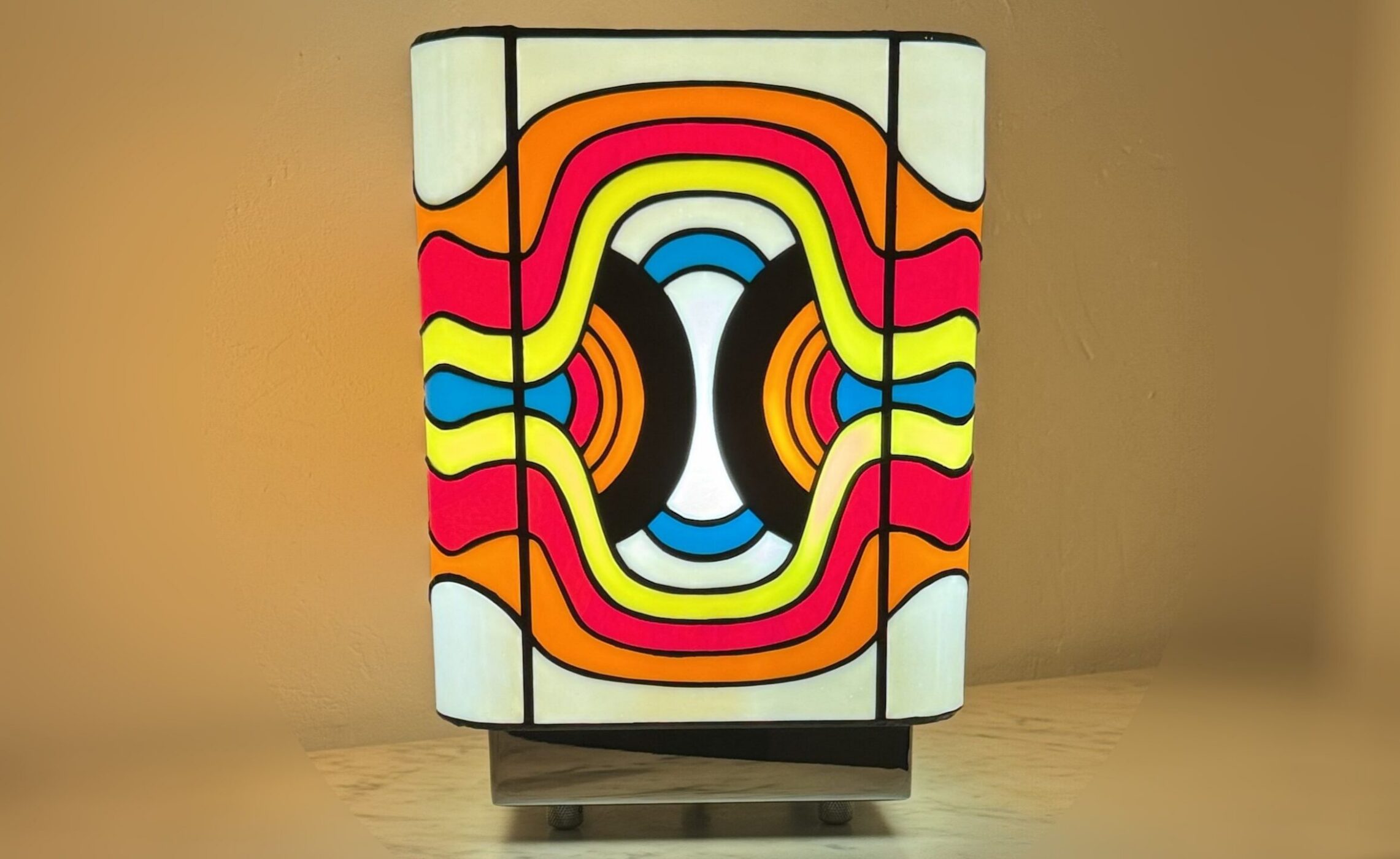 Lampe de table cubique en vitrail Tiffany, de style Pop Art ou Rétro-Futuriste (années 60-70). Chaque face de la lampe présente un motif psychédélique et abstrait composé de vagues, de lignes courbes et de formes ovales imbriquées. La palette de couleurs est extrêmement vive et contrastée : rouge vif, jaune citron, bleu turquoise, orange et noir, sur un fond blanc opalescent. Les soudures sont noires. La lampe repose sur une base claire (bois ou métal) et diffuse une lumière chaude, mettant en valeur son design dynamique et coloré.