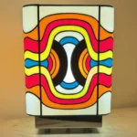 Lampe de table cubique en vitrail Tiffany, de style Pop Art ou Rétro-Futuriste (années 60-70). Chaque face de la lampe présente un motif psychédélique et abstrait composé de vagues, de lignes courbes et de formes ovales imbriquées. La palette de couleurs est extrêmement vive et contrastée : rouge vif, jaune citron, bleu turquoise, orange et noir, sur un fond blanc opalescent. Les soudures sont noires. La lampe repose sur une base claire (bois ou métal) et diffuse une lumière chaude, mettant en valeur son design dynamique et coloré.