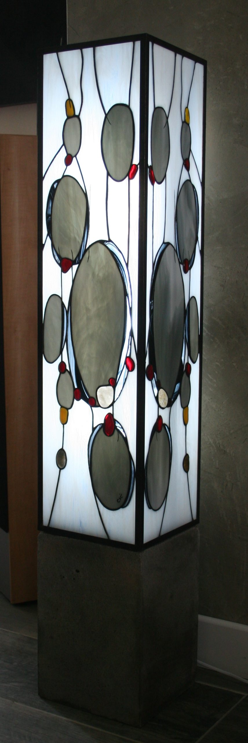 "Luminaire design en vitrail contemporain, colonne carrée aux motifs abstraits organiques : cercles gris translucides entrelacés de lignes noires, ponctués de touches rouges et dorées. Éclairage intérieur diffusant une lumière douce. Style moderne inspiré de l'art verrier.