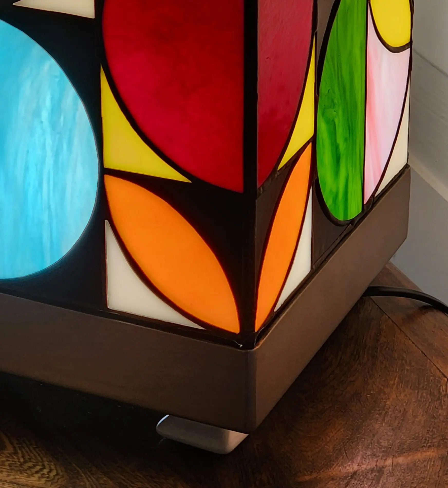 Lampe de table cubique de style Art Contemporain / Seventies, réalisée en vitrail Tiffany. La lampe est un cube en verre coloré, où chaque face présente un motif abstrait de cercles, demi-cercles, triangles et rectangles. La palette de couleurs est vibrante et multicolore, incluant du rouge, du bleu cobalt, du jaune citron, du vert forêt et de l'orange, le tout bordé de lignes de soudure noires ou cuivrées. Elle repose sur une base carrée inox, mettant en valeur son design géométrique et lumineux, inspiré du Rubik's Cube ou des motifs Bauhaus.