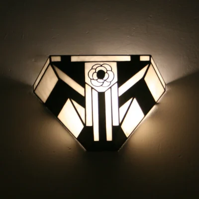 Applique murale Art Déco de style vitrail Tiffany, de forme pentagonale évasée. Elle présente un motif géométrique bicolore fort en noir et blanc opalescent. Le centre est orné d'une fleur stylisée (camélia) blanche et noire, encadrée par des bandes verticales noires et blanches. L'applique diffuse une lumière chaleureuse vers le haut et le bas, créant un éclairage d'ambiance contrasté.