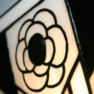 Applique murale Art Déco de style vitrail Tiffany, de forme pentagonale évasée. Elle présente un motif géométrique bicolore fort en noir et blanc opalescent. Le centre est orné d'une fleur stylisée (camélia) blanche et noire, encadrée par des bandes verticales noires et blanches. L'applique diffuse une lumière chaleureuse vers le haut et le bas, créant un éclairage d'ambiance contrasté.
