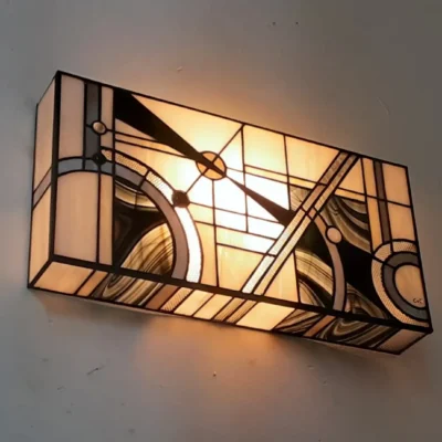 Applique murale rectangulaire de style Art Déco en vitrail Tiffany. Le motif abstrait et géométrique présente des lignes dynamiques, des cercles, et des triangles, évoquant un mouvement d'horlogerie ou de vitesse. La palette de couleurs est composée de blanc opalescent pour le fond, de noir mat, et de touches subtiles de gris ou de verre fumé marbré, créant un fort contraste. La lumière douce et chaude filtre à travers le panneau pour un éclairage d'accentuation.