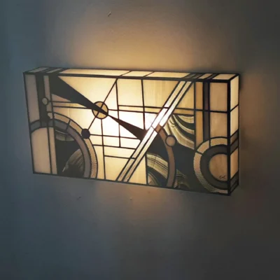 Applique murale rectangulaire de style Art Déco en vitrail Tiffany. Le motif abstrait et géométrique présente des lignes dynamiques, des cercles, et des triangles, évoquant un mouvement d'horlogerie ou de vitesse. La palette de couleurs est composée de blanc opalescent pour le fond, de noir mat, et de touches subtiles de gris ou de verre fumé marbré, créant un fort contraste. La lumière douce et chaude filtre à travers le panneau pour un éclairage d'accentuation.