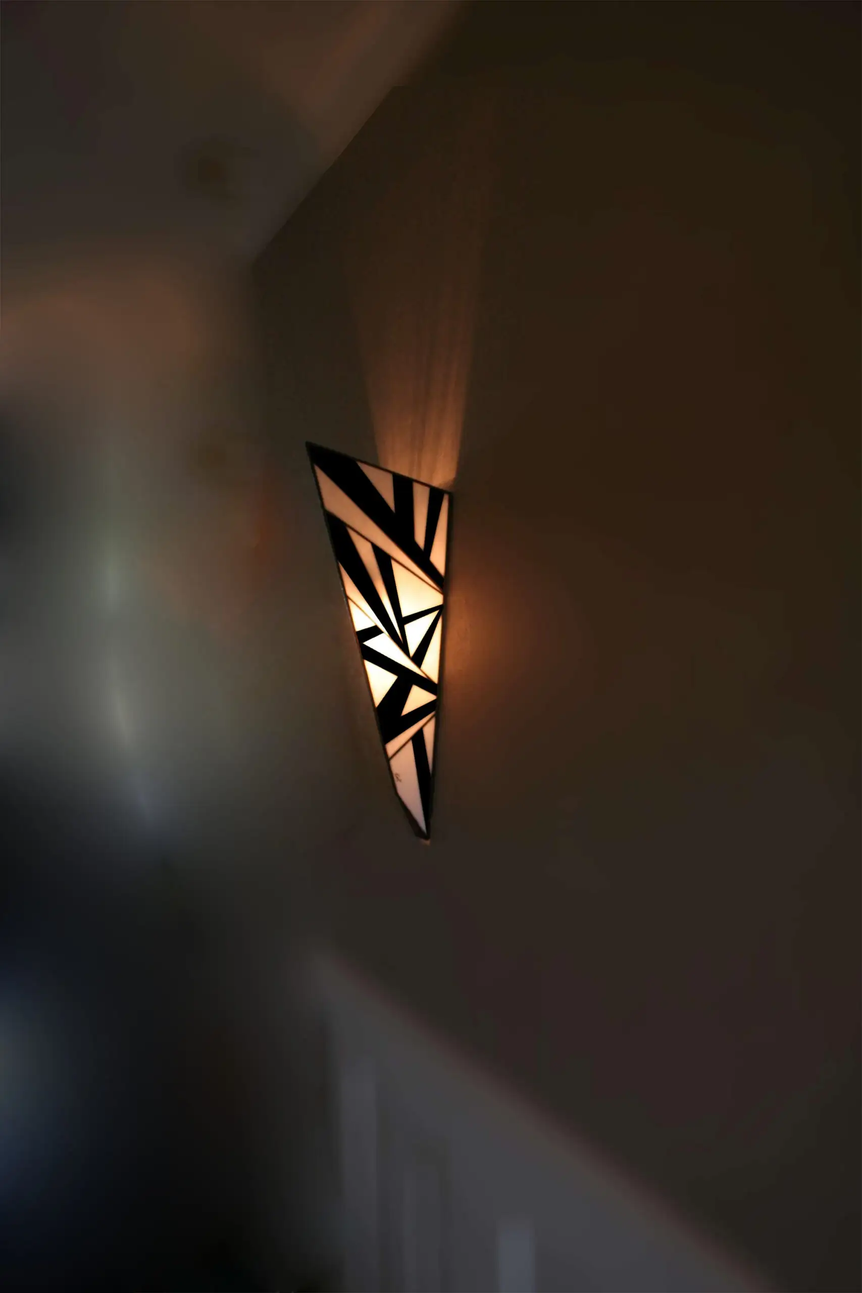 Applique murale verticale de style Art Déco en vitrail Tiffany, de forme effilée et conique. Le motif est composé de multiples triangles et formes angulaires, créant un design très dynamique et fracturé, presque abstrait. La palette de couleurs est un contraste fort et graphique entre le noir et le blanc opalescent. L'applique projette une lumière chaude vers le haut et vers le bas, accentuant son relief et son motif aiguisé.
