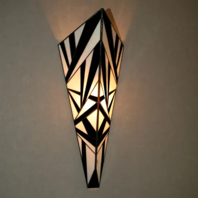 Applique murale verticale de style Art Déco en vitrail Tiffany, de forme effilée et conique. Le motif est composé de multiples triangles et formes angulaires, créant un design très dynamique et fracturé, presque abstrait. La palette de couleurs est un contraste fort et graphique entre le noir et le blanc opalescent. L'applique projette une lumière chaude vers le haut et vers le bas, accentuant son relief et son motif aiguisé.