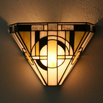 Applique murale Art Déco de style Tiffany, de forme trapézoïdale et en verre teinté. Le motif géométrique présente des lignes droites et des formes circulaires dans une palette de couleurs dominée par le blanc laiteux, le jaune ambre et le noir, avec des touches de verre texturé. La lumière chaude émise par l'applique crée un éclairage mural à la fois vers le haut et le bas, mettant en valeur son design géométrique et ses couleurs.