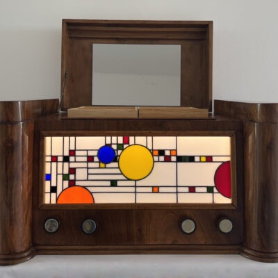 Coffre en bois foncé poli, de style Art Moderne, avec des formes arrondies sur les côtés. La façade principale est ornée d'un panneau de vitrail rétroéclairé. Le motif du vitrail est abstrait et géométrique, inspiré par le mouvement De Stijl (Piet Mondrian), composé de rectangles et de lignes noires sur fond blanc opalescent, intégrant des cercles colorés (jaune, bleu, orange, rouge). Le coffre possède deux tiroirs cachés sur les côtés et un couvercle supérieur qui s'ouvre pour révéler un miroir et un compartiment. Trois boutons ronds sont alignés sous le vitrail. L'éclairage LED interne diffuse une lumière douce à travers le vitrail.