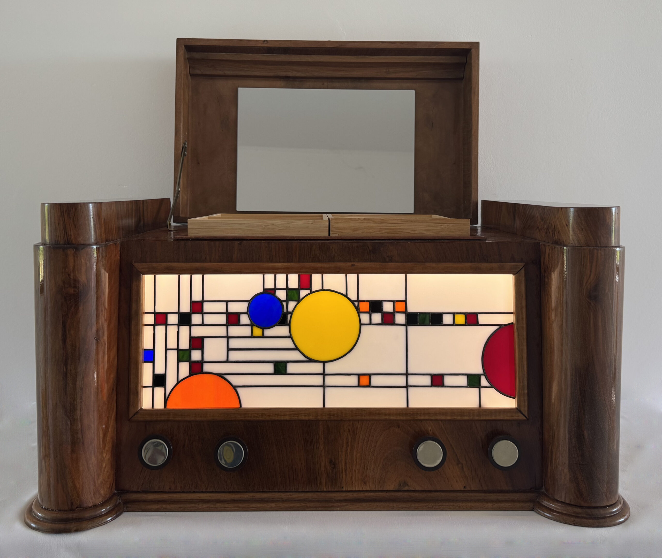 Coffre en bois foncé poli, de style Art Moderne, avec des formes arrondies sur les côtés. La façade principale est ornée d'un panneau de vitrail rétroéclairé. Le motif du vitrail est abstrait et géométrique, inspiré par le mouvement De Stijl (Piet Mondrian), composé de rectangles et de lignes noires sur fond blanc opalescent, intégrant des cercles colorés (jaune, bleu, orange, rouge). Le coffre possède deux tiroirs cachés sur les côtés et un couvercle supérieur qui s'ouvre pour révéler un miroir et un compartiment. Trois boutons ronds sont alignés sous le vitrail. L'éclairage LED interne diffuse une lumière douce à travers le vitrail.