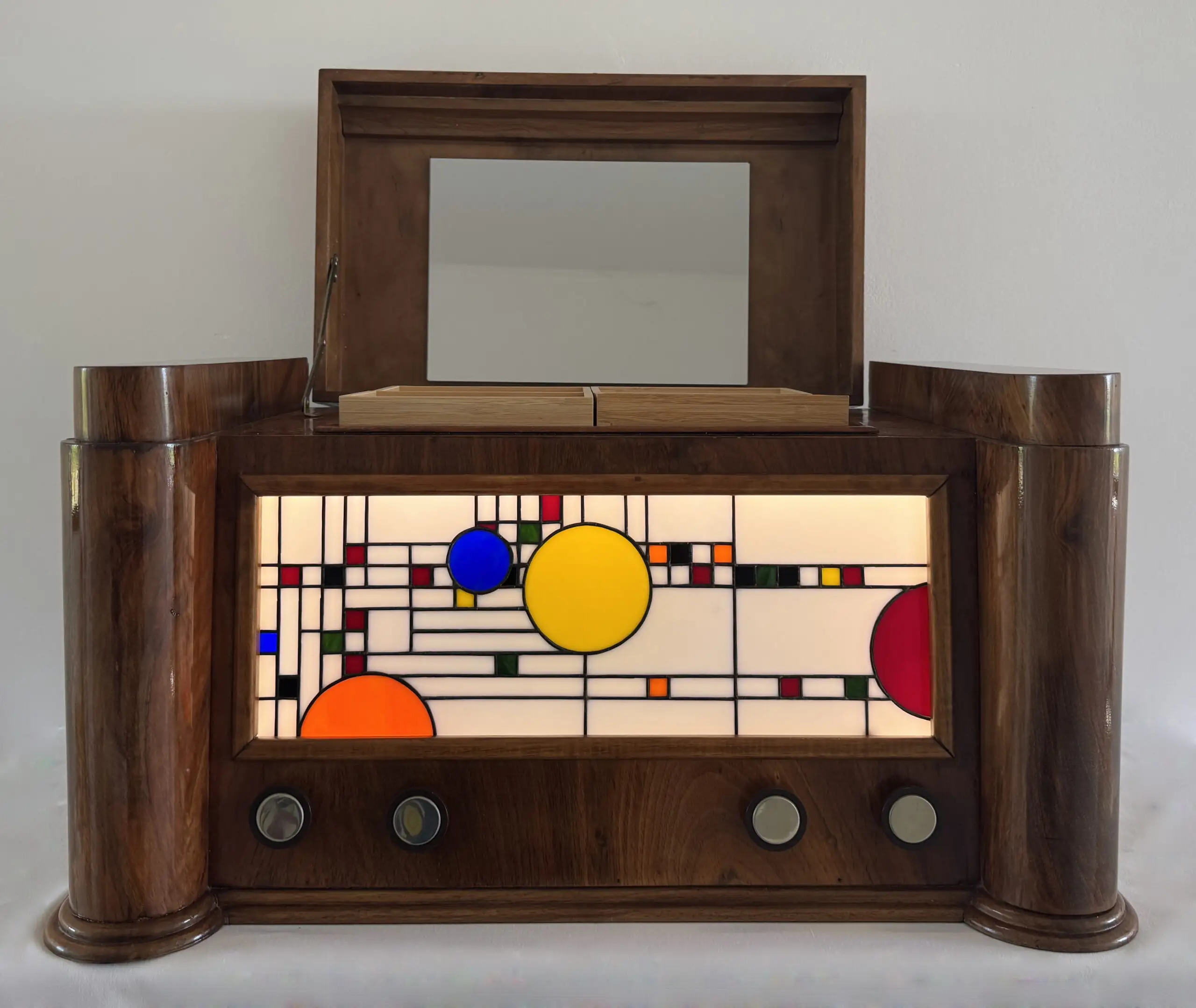 Coffre en bois foncé poli, de style Art Moderne, avec des formes arrondies sur les côtés. La façade principale est ornée d'un panneau de vitrail rétroéclairé. Le motif du vitrail est abstrait et géométrique, inspiré par le mouvement De Stijl (Piet Mondrian), composé de rectangles et de lignes noires sur fond blanc opalescent, intégrant des cercles colorés (jaune, bleu, orange, rouge). Le coffre possède deux tiroirs cachés sur les côtés et un couvercle supérieur qui s'ouvre pour révéler un miroir et un compartiment. Trois boutons ronds sont alignés sous le vitrail. L'éclairage LED interne diffuse une lumière douce à travers le vitrail.
