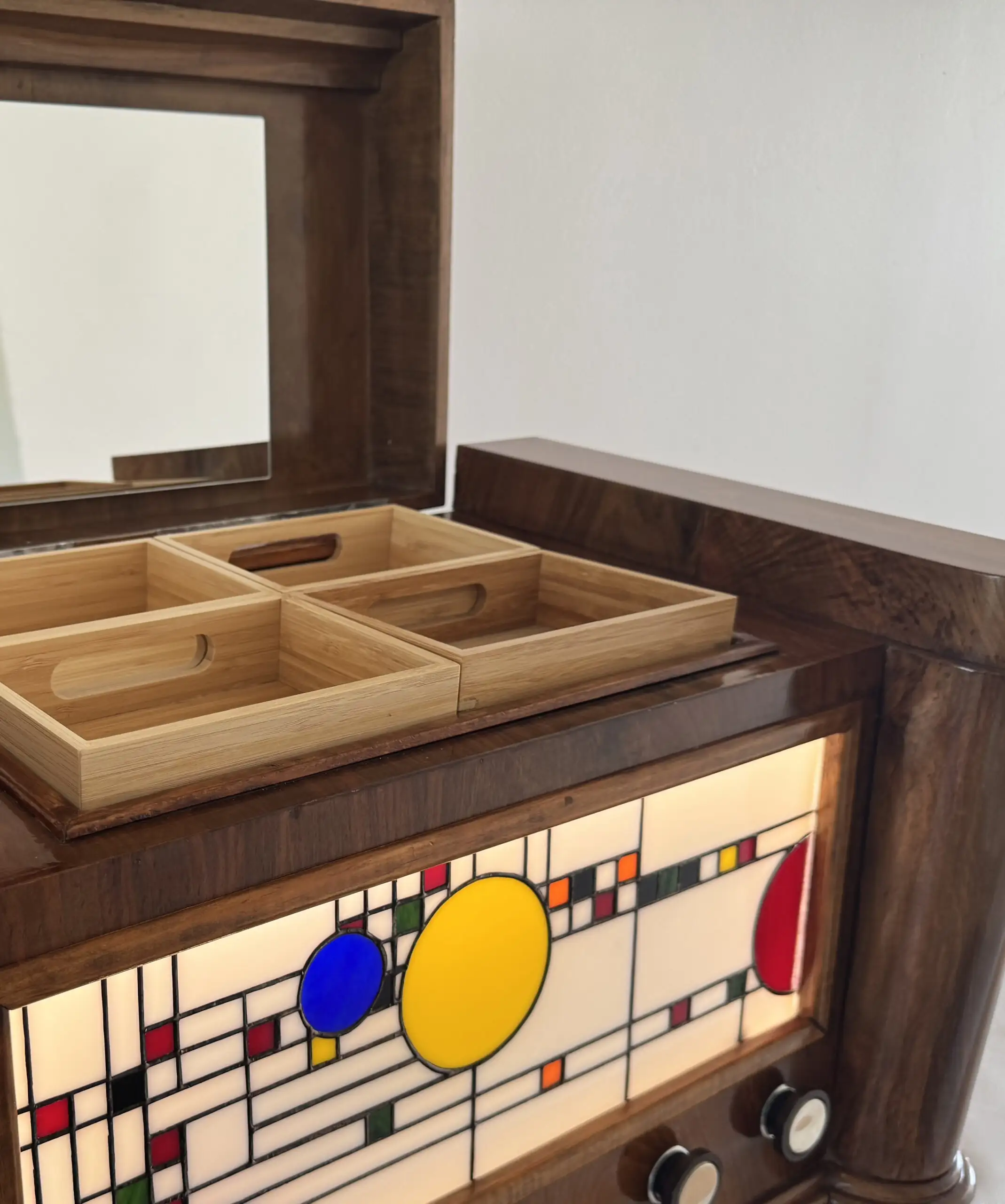 Coffre en bois foncé poli, de style Art Moderne, avec des formes arrondies sur les côtés. La façade principale est ornée d'un panneau de vitrail rétroéclairé. Le motif du vitrail est abstrait et géométrique, inspiré par le mouvement De Stijl (Piet Mondrian), composé de rectangles et de lignes noires sur fond blanc opalescent, intégrant des cercles colorés (jaune, bleu, orange, rouge). Le coffre possède deux tiroirs cachés sur les côtés et un couvercle supérieur qui s'ouvre pour révéler un miroir et un compartiment. Trois boutons ronds sont alignés sous le vitrail. L'éclairage LED interne diffuse une lumière douce à travers le vitrail.