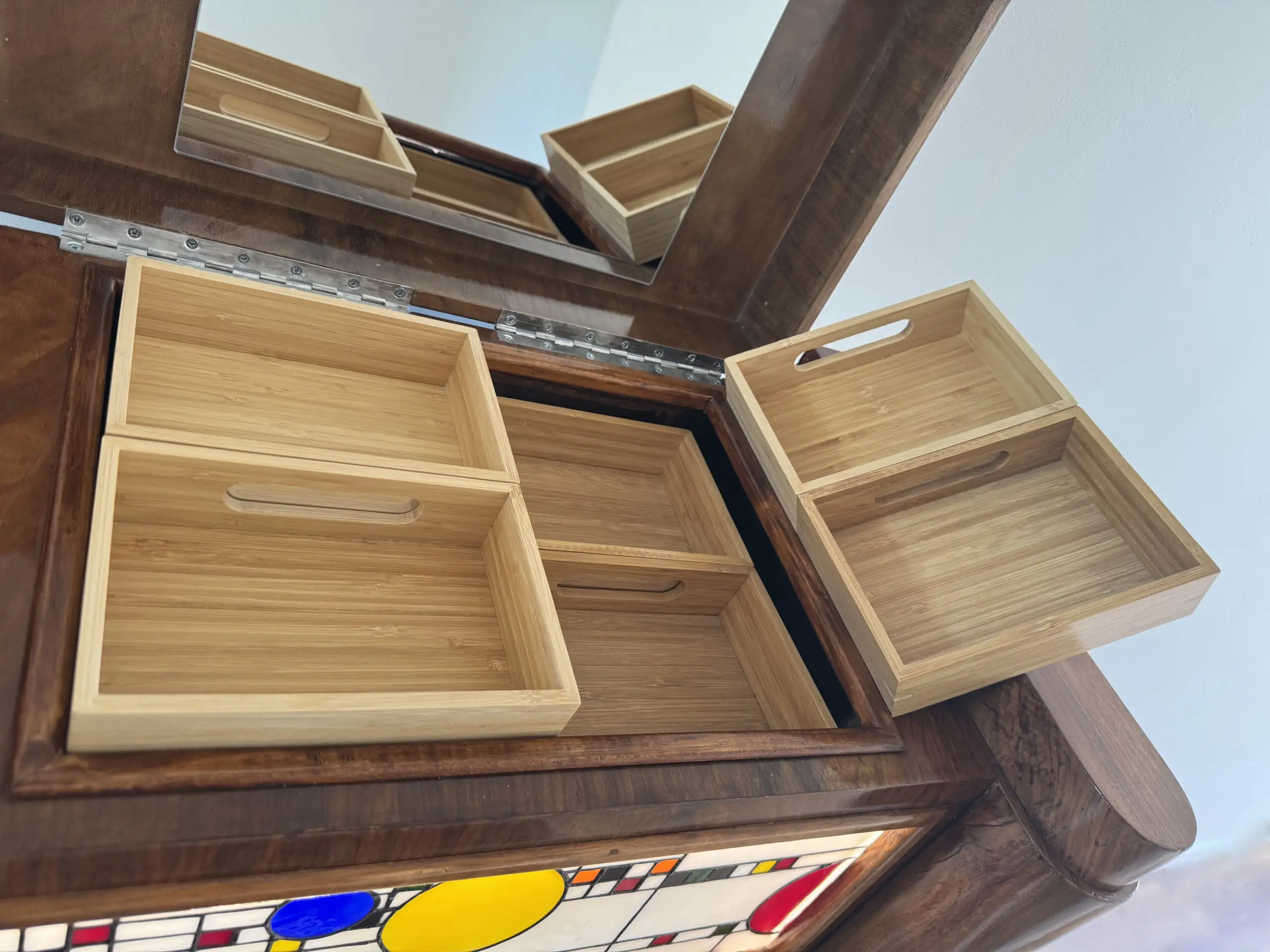 Coffre en bois foncé poli, de style Art Moderne, avec des formes arrondies sur les côtés. La façade principale est ornée d'un panneau de vitrail rétroéclairé. Le motif du vitrail est abstrait et géométrique, inspiré par le mouvement De Stijl (Piet Mondrian), composé de rectangles et de lignes noires sur fond blanc opalescent, intégrant des cercles colorés (jaune, bleu, orange, rouge). Le coffre possède deux tiroirs cachés sur les côtés et un couvercle supérieur qui s'ouvre pour révéler un miroir et un compartiment. Trois boutons ronds sont alignés sous le vitrail. L'éclairage LED interne diffuse une lumière douce à travers le vitrail.