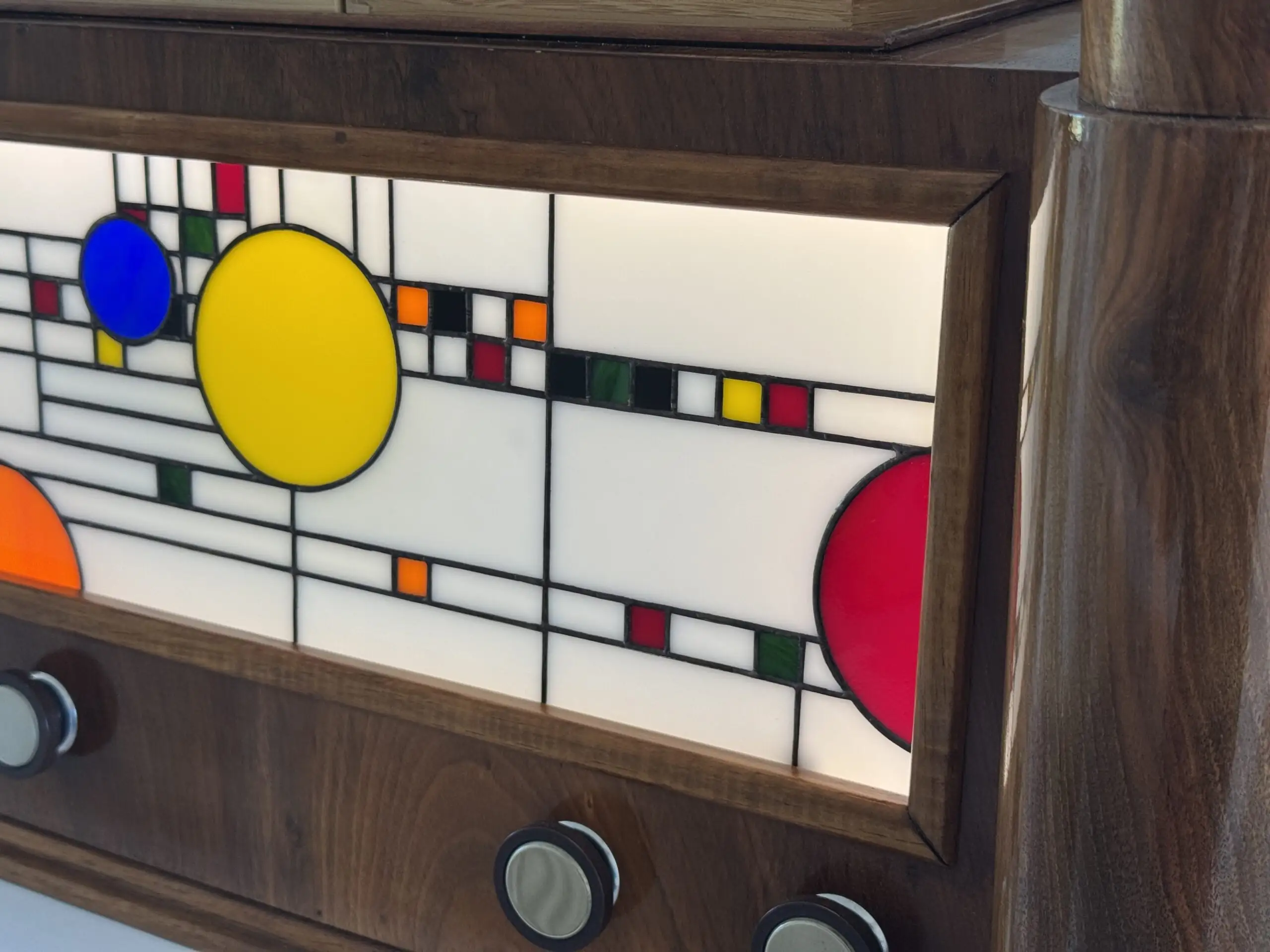 Coffre en bois foncé poli, de style Art Moderne, avec des formes arrondies sur les côtés. La façade principale est ornée d'un panneau de vitrail rétroéclairé. Le motif du vitrail est abstrait et géométrique, inspiré par le mouvement De Stijl (Piet Mondrian), composé de rectangles et de lignes noires sur fond blanc opalescent, intégrant des cercles colorés (jaune, bleu, orange, rouge). Le coffre possède deux tiroirs cachés sur les côtés et un couvercle supérieur qui s'ouvre pour révéler un miroir et un compartiment. Trois boutons ronds sont alignés sous le vitrail. L'éclairage LED interne diffuse une lumière douce à travers le vitrail.