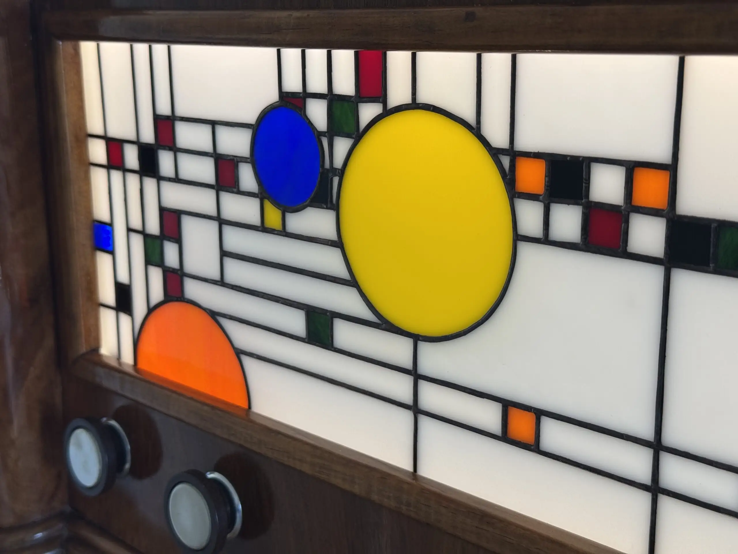 Coffre en bois foncé poli, de style Art Moderne, avec des formes arrondies sur les côtés. La façade principale est ornée d'un panneau de vitrail rétroéclairé. Le motif du vitrail est abstrait et géométrique, inspiré par le mouvement De Stijl (Piet Mondrian), composé de rectangles et de lignes noires sur fond blanc opalescent, intégrant des cercles colorés (jaune, bleu, orange, rouge). Le coffre possède deux tiroirs cachés sur les côtés et un couvercle supérieur qui s'ouvre pour révéler un miroir et un compartiment. Trois boutons ronds sont alignés sous le vitrail. L'éclairage LED interne diffuse une lumière douce à travers le vitrail.