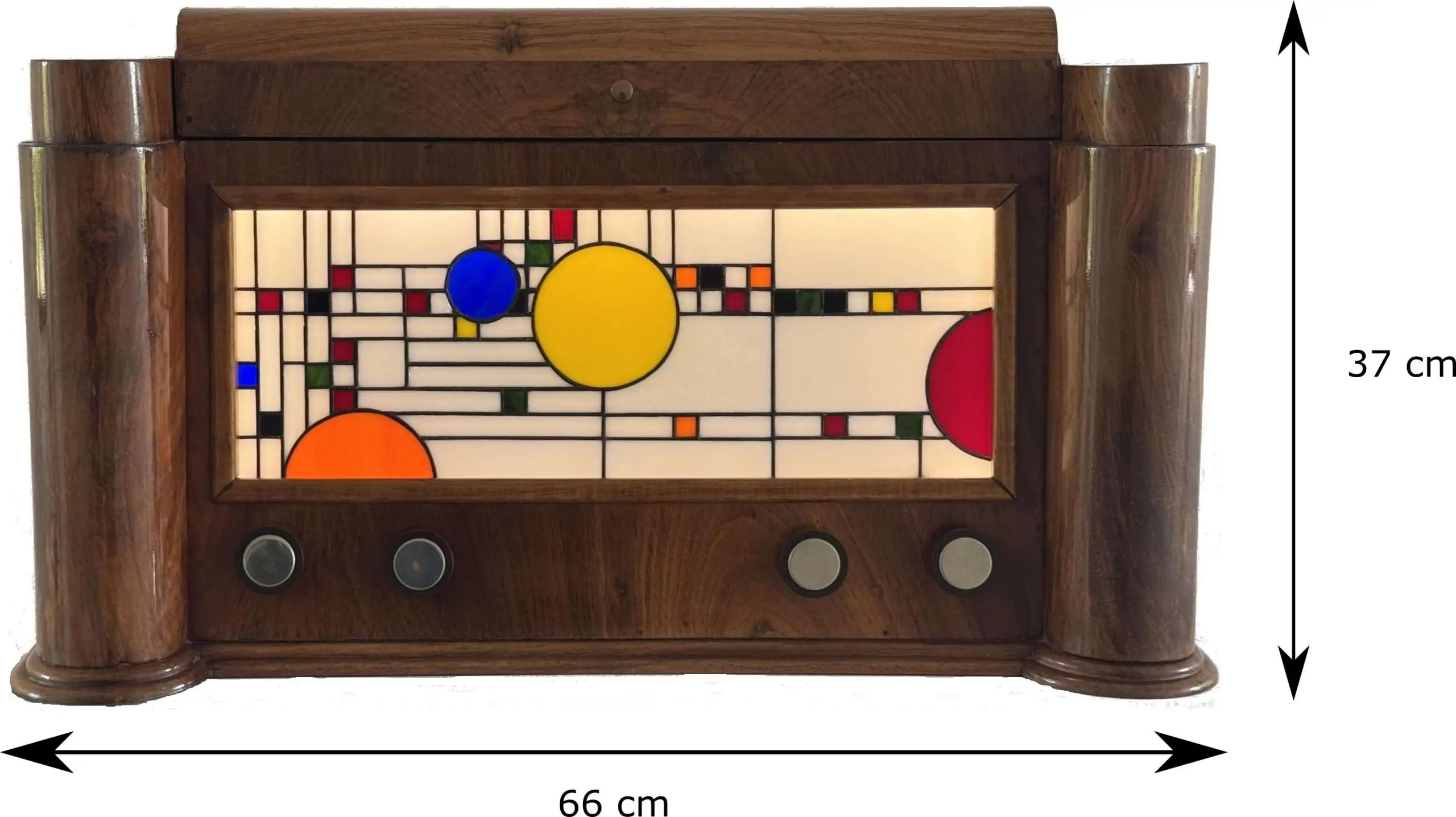 Coffre en bois foncé poli, de style Art Moderne, avec des formes arrondies sur les côtés. La façade principale est ornée d'un panneau de vitrail rétroéclairé. Le motif du vitrail est abstrait et géométrique, inspiré par le mouvement De Stijl (Piet Mondrian), composé de rectangles et de lignes noires sur fond blanc opalescent, intégrant des cercles colorés (jaune, bleu, orange, rouge). Le coffre possède deux tiroirs cachés sur les côtés et un couvercle supérieur qui s'ouvre pour révéler un miroir et un compartiment. Trois boutons ronds sont alignés sous le vitrail. L'éclairage LED interne diffuse une lumière douce à travers le vitrail.