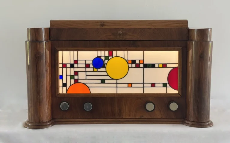 Coffre en bois foncé poli, de style Art Moderne, avec des formes arrondies sur les côtés. La façade principale est ornée d'un panneau de vitrail rétroéclairé. Le motif du vitrail est abstrait et géométrique, inspiré par le mouvement De Stijl (Piet Mondrian), composé de rectangles et de lignes noires sur fond blanc opalescent, intégrant des cercles colorés (jaune, bleu, orange, rouge). Le coffre possède deux tiroirs cachés sur les côtés et un couvercle supérieur qui s'ouvre pour révéler un miroir et un compartiment. Trois boutons ronds sont alignés sous le vitrail. L'éclairage LED interne diffuse une lumière douce à travers le vitrail.