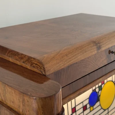 Coffre en bois foncé poli, de style Art Moderne, avec des formes arrondies sur les côtés. La façade principale est ornée d'un panneau de vitrail rétroéclairé. Le motif du vitrail est abstrait et géométrique, inspiré par le mouvement De Stijl (Piet Mondrian), composé de rectangles et de lignes noires sur fond blanc opalescent, intégrant des cercles colorés (jaune, bleu, orange, rouge). Le coffre possède deux tiroirs cachés sur les côtés et un couvercle supérieur qui s'ouvre pour révéler un miroir et un compartiment. Trois boutons ronds sont alignés sous le vitrail. L'éclairage LED interne diffuse une lumière douce à travers le vitrail.