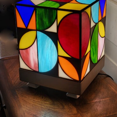 Lampe de table cubique de style Art Contemporain / Seventies, réalisée en vitrail Tiffany. La lampe est un cube en verre coloré, où chaque face présente un motif abstrait de cercles, demi-cercles, triangles et rectangles. La palette de couleurs est vibrante et multicolore, incluant du rouge, du bleu cobalt, du jaune citron, du vert forêt et de l'orange, le tout bordé de lignes de soudure noires ou cuivrées. Elle repose sur une base carrée inox, mettant en valeur son design géométrique et lumineux, inspiré du Rubik's Cube ou des motifs Bauhaus.