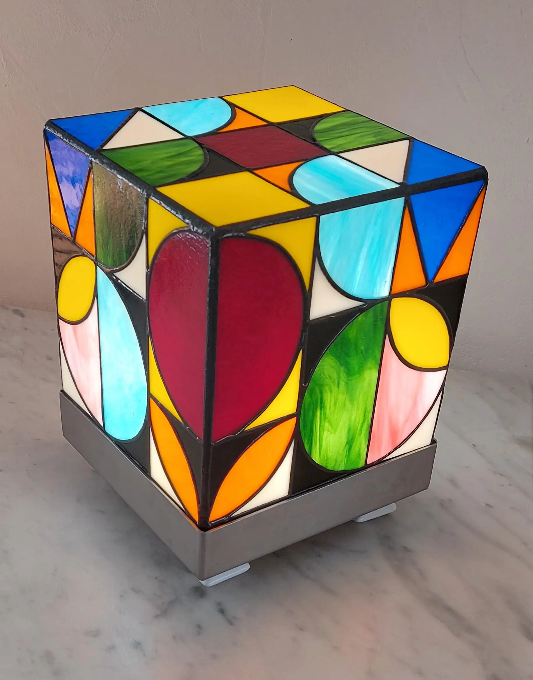 Lampe de table cubique de style Art Contemporain / Seventies, réalisée en vitrail Tiffany. La lampe est un cube en verre coloré, où chaque face présente un motif abstrait de cercles, demi-cercles, triangles et rectangles. La palette de couleurs est vibrante et multicolore, incluant du rouge, du bleu cobalt, du jaune citron, du vert forêt et de l'orange, le tout bordé de lignes de soudure noires ou cuivrées. Elle repose sur une base carrée inox, mettant en valeur son design géométrique et lumineux, inspiré du Rubik's Cube ou des motifs Bauhaus.
