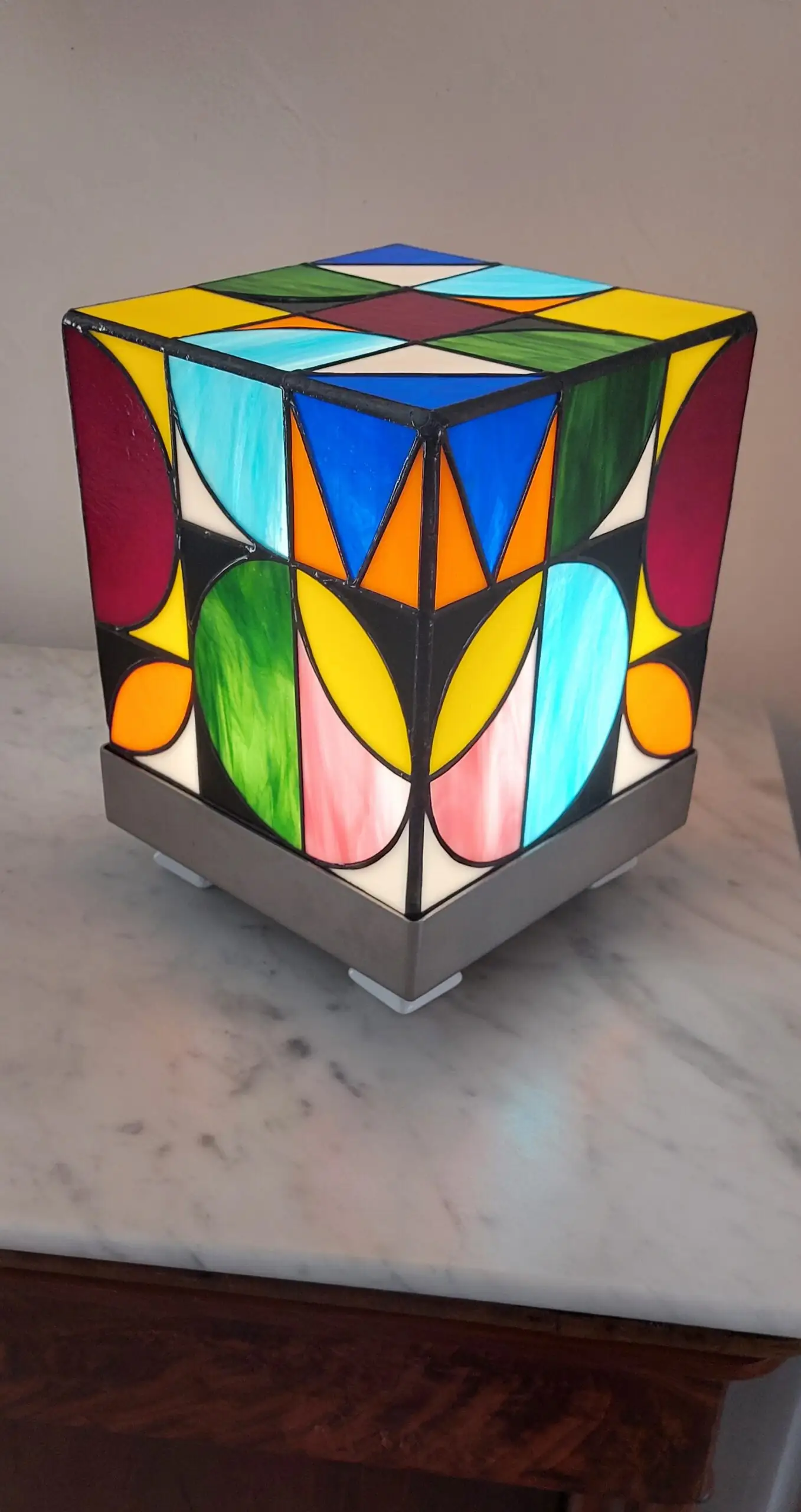 Lampe de table cubique de style Art Contemporain / Seventies, réalisée en vitrail Tiffany. La lampe est un cube en verre coloré, où chaque face présente un motif abstrait de cercles, demi-cercles, triangles et rectangles. La palette de couleurs est vibrante et multicolore, incluant du rouge, du bleu cobalt, du jaune citron, du vert forêt et de l'orange, le tout bordé de lignes de soudure noires ou cuivrées. Elle repose sur une base carrée inox, mettant en valeur son design géométrique et lumineux, inspiré du Rubik's Cube ou des motifs Bauhaus.