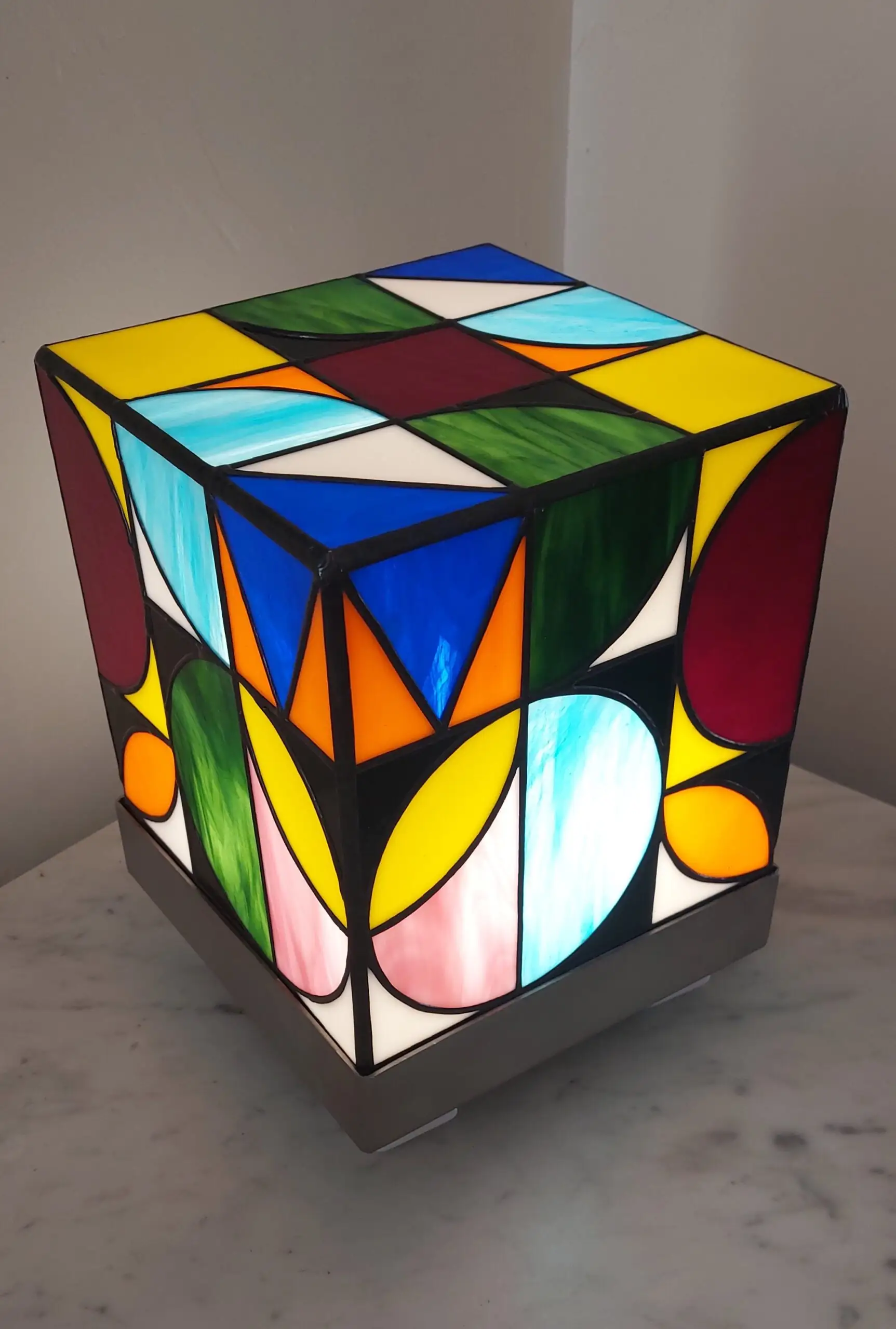 Lampe de table cubique de style Art Contemporain / Seventies, réalisée en vitrail Tiffany. La lampe est un cube en verre coloré, où chaque face présente un motif abstrait de cercles, demi-cercles, triangles et rectangles. La palette de couleurs est vibrante et multicolore, incluant du rouge, du bleu cobalt, du jaune citron, du vert forêt et de l'orange, le tout bordé de lignes de soudure noires ou cuivrées. Elle repose sur une base carrée inox, mettant en valeur son design géométrique et lumineux, inspiré du Rubik's Cube ou des motifs Bauhaus.