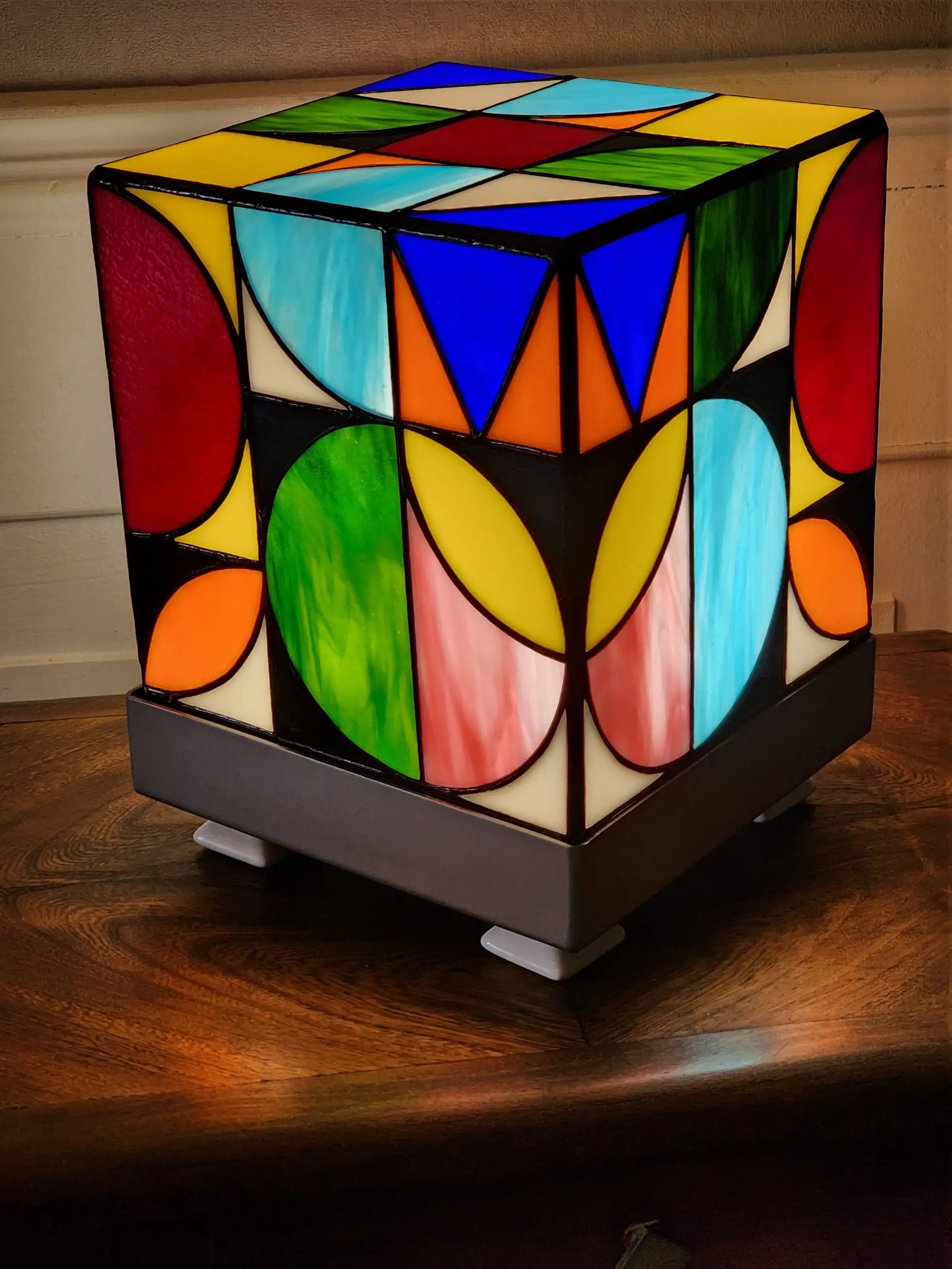 Lampe de table cubique de style Art Contemporain / Seventies, réalisée en vitrail Tiffany. La lampe est un cube en verre coloré, où chaque face présente un motif abstrait de cercles, demi-cercles, triangles et rectangles. La palette de couleurs est vibrante et multicolore, incluant du rouge, du bleu cobalt, du jaune citron, du vert forêt et de l'orange, le tout bordé de lignes de soudure noires ou cuivrées. Elle repose sur une base carrée inox, mettant en valeur son design géométrique et lumineux, inspiré du Rubik's Cube ou des motifs Bauhaus.