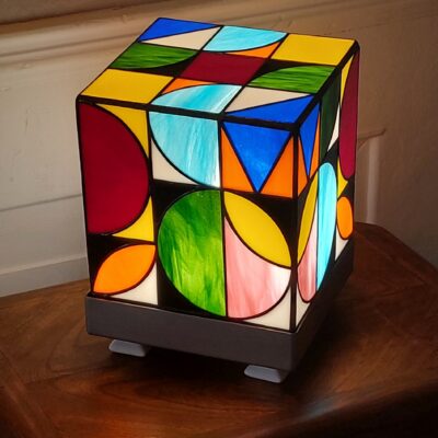 Lampe de table cubique de style Art Contemporain / Seventies, réalisée en vitrail Tiffany. La lampe est un cube en verre coloré, où chaque face présente un motif abstrait de cercles, demi-cercles, triangles et rectangles. La palette de couleurs est vibrante et multicolore, incluant du rouge, du bleu cobalt, du jaune citron, du vert forêt et de l'orange, le tout bordé de lignes de soudure noires ou cuivrées. Elle repose sur une base carrée inox, mettant en valeur son design géométrique et lumineux, inspiré du Rubik's Cube ou des motifs Bauhaus.