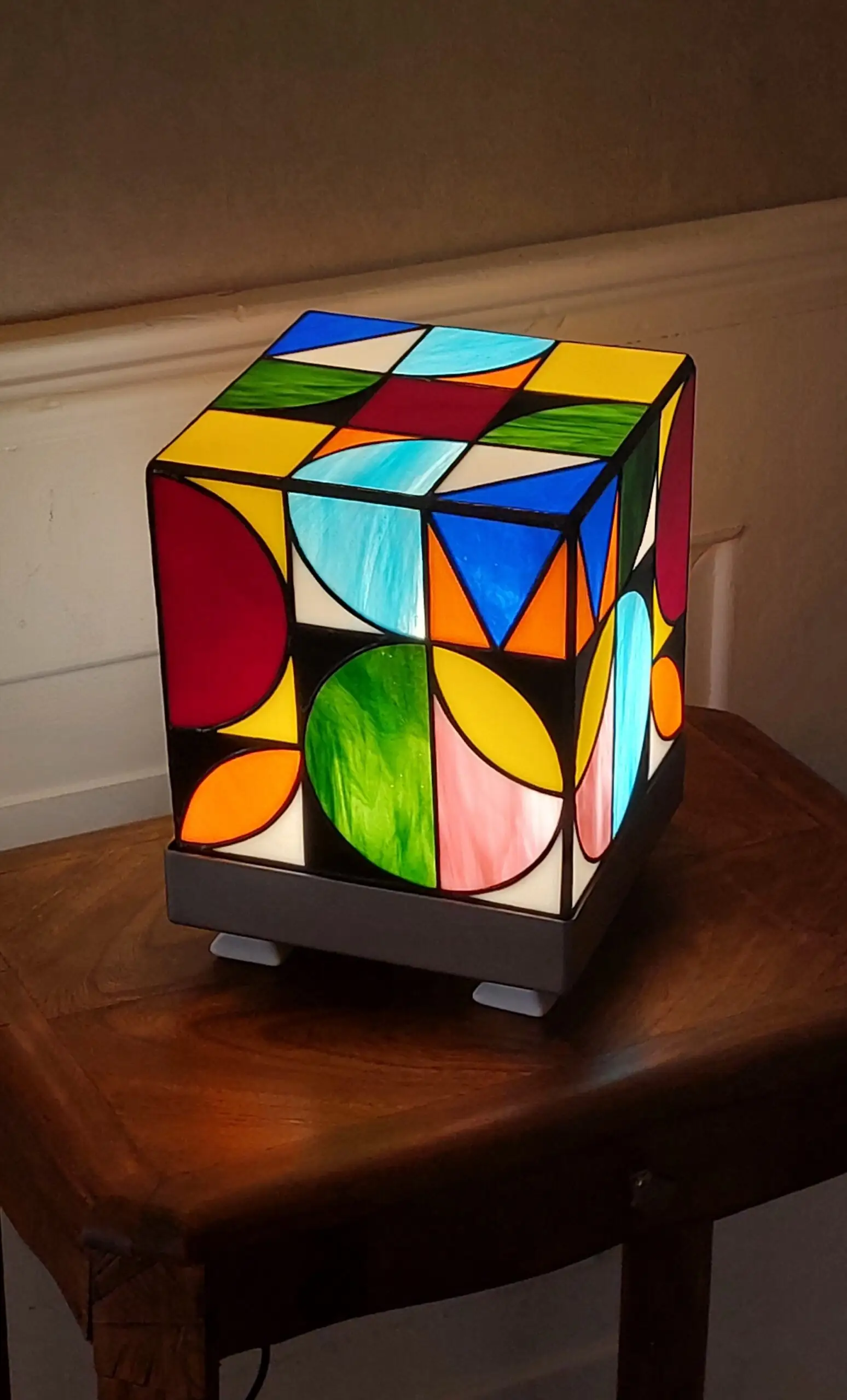 Lampe de table cubique de style Art Contemporain / Seventies, réalisée en vitrail Tiffany. La lampe est un cube en verre coloré, où chaque face présente un motif abstrait de cercles, demi-cercles, triangles et rectangles. La palette de couleurs est vibrante et multicolore, incluant du rouge, du bleu cobalt, du jaune citron, du vert forêt et de l'orange, le tout bordé de lignes de soudure noires ou cuivrées. Elle repose sur une base carrée inox, mettant en valeur son design géométrique et lumineux, inspiré du Rubik's Cube ou des motifs Bauhaus.