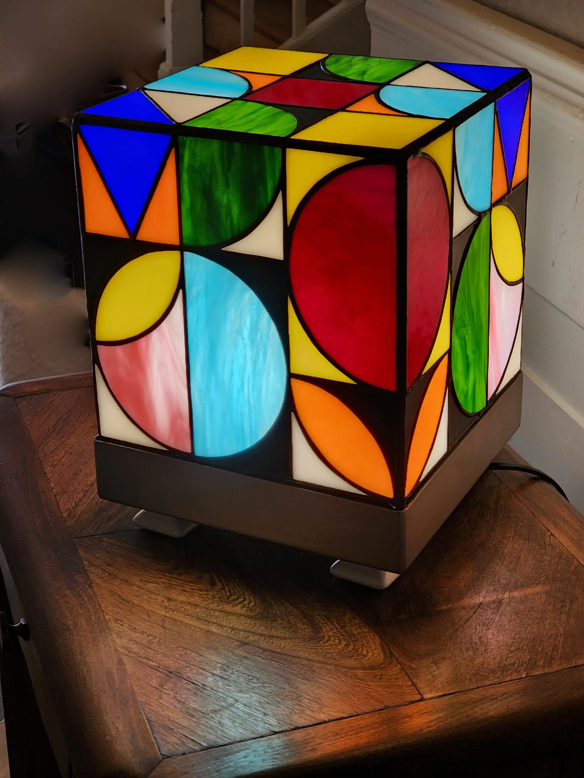 Lampe de table cubique de style Art Contemporain / Seventies, réalisée en vitrail Tiffany. La lampe est un cube en verre coloré, où chaque face présente un motif abstrait de cercles, demi-cercles, triangles et rectangles. La palette de couleurs est vibrante et multicolore, incluant du rouge, du bleu cobalt, du jaune citron, du vert forêt et de l'orange, le tout bordé de lignes de soudure noires ou cuivrées. Elle repose sur une base carrée inox, mettant en valeur son design géométrique et lumineux, inspiré du Rubik's Cube ou des motifs Bauhaus.