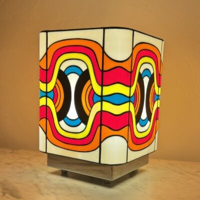 Lampe de table cubique en vitrail Tiffany, de style Pop Art ou Rétro-Futuriste (années 60-70). Chaque face de la lampe présente un motif psychédélique et abstrait composé de vagues, de lignes courbes et de formes ovales imbriquées. La palette de couleurs est extrêmement vive et contrastée : rouge vif, jaune citron, bleu turquoise, orange et noir, sur un fond blanc opalescent. Les soudures sont noires. La lampe repose sur une base claire (bois ou métal) et diffuse une lumière chaude, mettant en valeur son design dynamique et coloré.