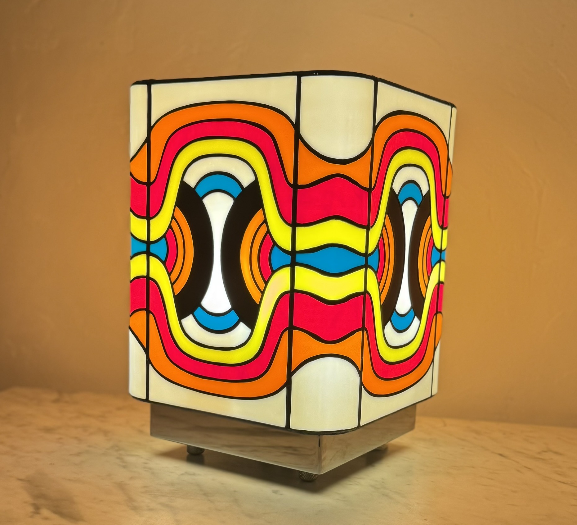 Lampe de table cubique en vitrail Tiffany, de style Pop Art ou Rétro-Futuriste (années 60-70). Chaque face de la lampe présente un motif psychédélique et abstrait composé de vagues, de lignes courbes et de formes ovales imbriquées. La palette de couleurs est extrêmement vive et contrastée : rouge vif, jaune citron, bleu turquoise, orange et noir, sur un fond blanc opalescent. Les soudures sont noires. La lampe repose sur une base claire (bois ou métal) et diffuse une lumière chaude, mettant en valeur son design dynamique et coloré.