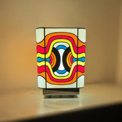 Lampe de table cubique en vitrail Tiffany, de style Pop Art ou Rétro-Futuriste (années 60-70). Chaque face de la lampe présente un motif psychédélique et abstrait composé de vagues, de lignes courbes et de formes ovales imbriquées. La palette de couleurs est extrêmement vive et contrastée : rouge vif, jaune citron, bleu turquoise, orange et noir, sur un fond blanc opalescent. Les soudures sont noires. La lampe repose sur une base claire (bois ou métal) et diffuse une lumière chaude, mettant en valeur son design dynamique et coloré.