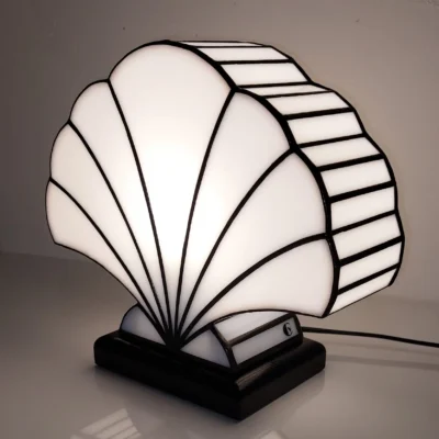 Lampe de table Art Déco en vitrail Tiffany, de forme coquillage stylisé. Le design est composé de panneaux de verre blanc opalescent et de soudures noires, créant une structure graphique et évasée. La base est noire, et la lampe est illuminée de l'intérieur, diffusant une lumière douce et uniforme qui met en valeur la forme et les lignes noires. Son reflet clair est visible sur la surface brillante sous la lampe, accentuant son élégance classique inspirée des années 1920-1930.