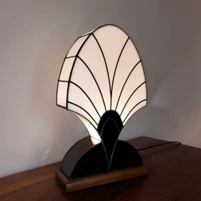 Lampe de table Art Déco en vitrail Tiffany, de forme haute et arrondie rappelant une palmette, un éventail fermé ou une plume stylisée. Le vitrail supérieur est en verre blanc opalescent, segmenté par des lignes radiales noires. La base incurvée est en verre noir opaque, intégrant un motif semi-circulaire accentué par un détail doré. L'ensemble repose sur un socle en bois, et la lumière douce filtre à travers le verre blanc, mettant en évidence le contraste spectaculaire entre le noir, le blanc et l'or.