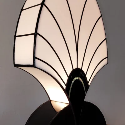 Lampe de table Art Déco en vitrail Tiffany, de forme haute et arrondie rappelant une palmette, un éventail fermé ou une plume stylisée. Le vitrail supérieur est en verre blanc opalescent, segmenté par des lignes radiales noires. La base incurvée est en verre noir opaque, intégrant un motif semi-circulaire accentué par un détail doré. L'ensemble repose sur un socle en bois, et la lumière douce filtre à travers le verre blanc, mettant en évidence le contraste spectaculaire entre le noir, le blanc et l'or.