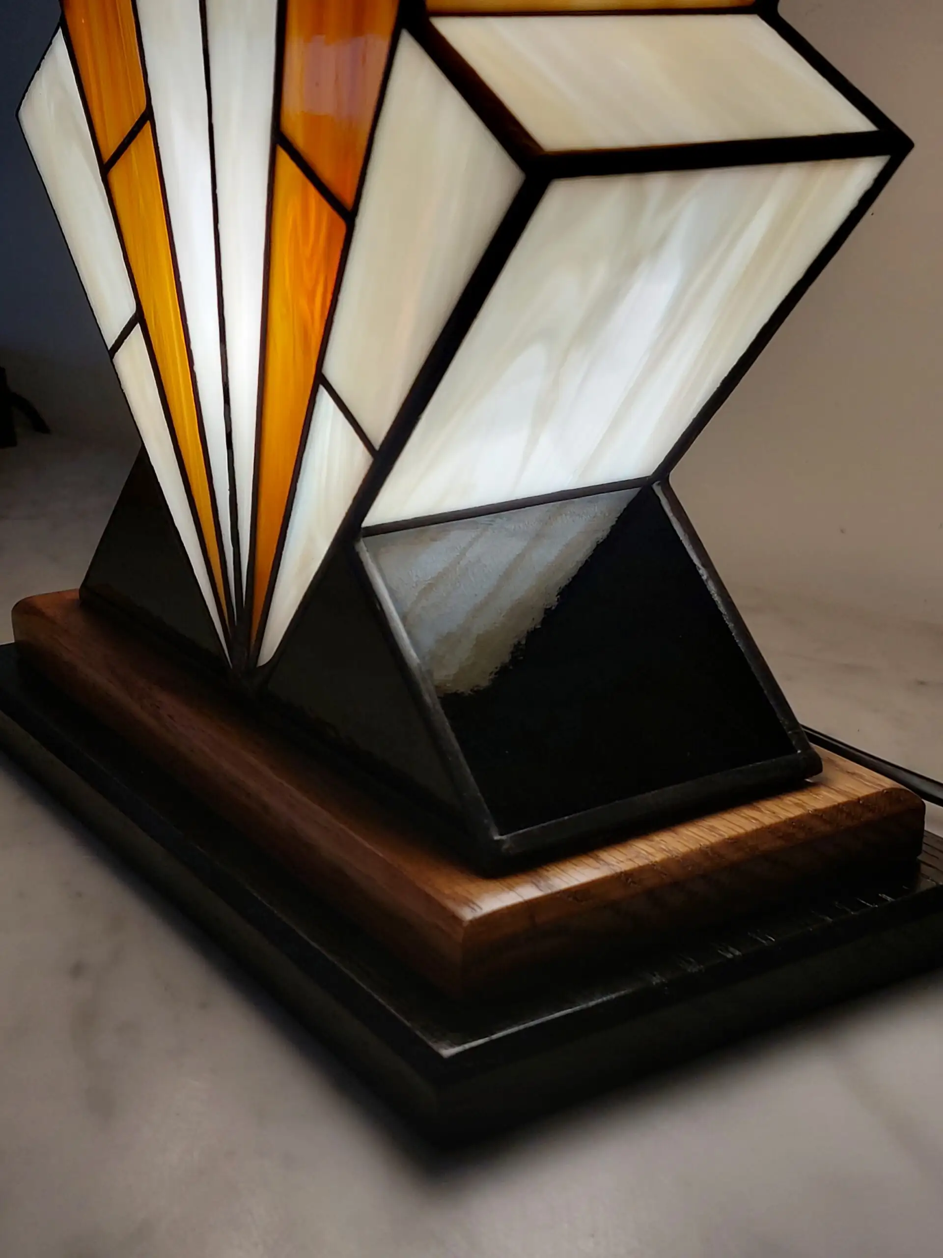 Grande lampe de table Art Déco en vitrail, de forme spectaculaire et évasée, évoquant la silhouette d'un gratte-ciel. L'abat-jour est composé de panneaux de verre ivoire Opalescent et de verre Ambre, alternant pour créer un effet de contraste vibrant. Les segments sont séparés par des soudures noires fines. Elle repose sur une base étagée en bois foncé et noir, et diffuse une lumière chaude et colorée, mettant en évidence son architecture géométrique et ses couleurs.