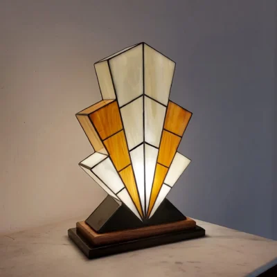 Grande lampe de table Art Déco en vitrail, de forme spectaculaire et évasée, évoquant la silhouette d'un gratte-ciel. L'abat-jour est composé de panneaux de verre ivoire Opalescent et de verre Ambre, alternant pour créer un effet de contraste vibrant. Les segments sont séparés par des soudures noires fines. Elle repose sur une base étagée en bois foncé et noir, et diffuse une lumière chaude et colorée, mettant en évidence son architecture géométrique et ses couleurs.