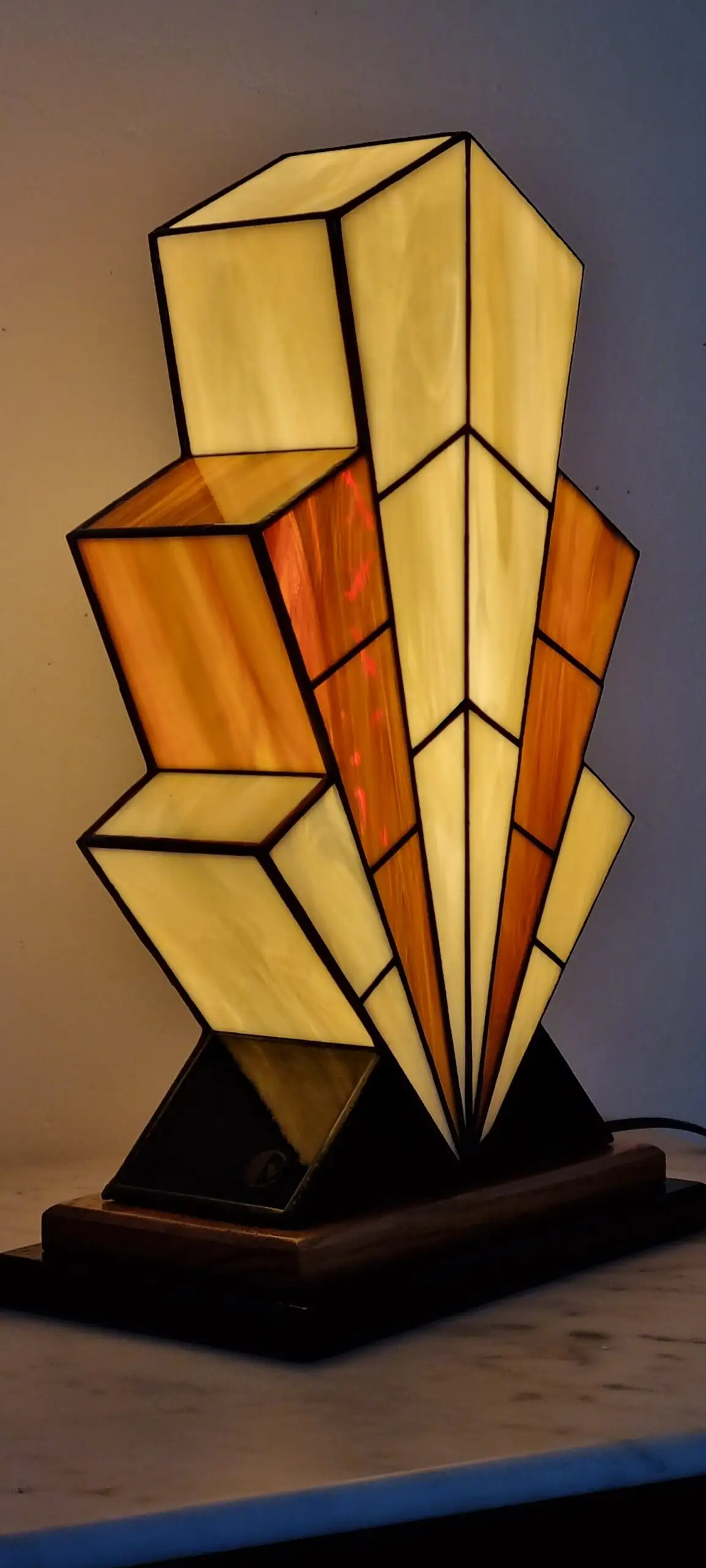 Grande lampe de table Art Déco en vitrail, de forme spectaculaire et évasée, évoquant la silhouette d'un gratte-ciel. L'abat-jour est composé de panneaux de verre ivoire Opalescent et de verre Ambre, alternant pour créer un effet de contraste vibrant. Les segments sont séparés par des soudures noires fines. Elle repose sur une base étagée en bois foncé et noir, et diffuse une lumière chaude et colorée, mettant en évidence son architecture géométrique et ses couleurs.