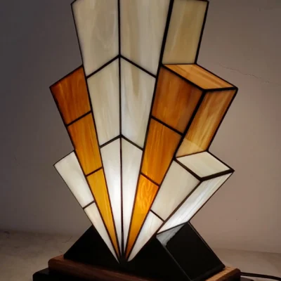 Grande lampe de table Art Déco en vitrail, de forme spectaculaire et évasée, évoquant la silhouette d'un gratte-ciel. L'abat-jour est composé de panneaux de verre ivoire Opalescent et de verre Ambre, alternant pour créer un effet de contraste vibrant. Les segments sont séparés par des soudures noires fines. Elle repose sur une base étagée en bois foncé et noir, et diffuse une lumière chaude et colorée, mettant en évidence son architecture géométrique et ses couleurs.