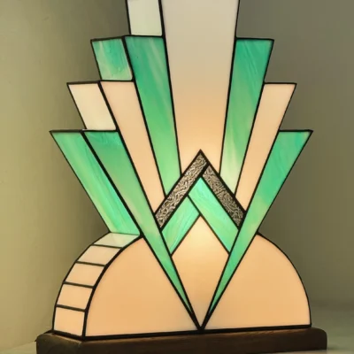 Lampe Art Déco en vitrail, inspiration Tiffany. Elle présente une base semi-circulaire blanche et un motif rayonnant et étagé de triangles vert-menthe et blancs, rappelant un gratte-ciel ou un éventail stylisé, avec une bande centrale argentée texturée.