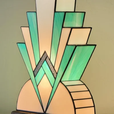 Lampe Art Déco en vitrail, inspiration Tiffany. Elle présente une base semi-circulaire blanche et un motif rayonnant et étagé de triangles vert-menthe et blancs, rappelant un gratte-ciel ou un éventail stylisé, avec une bande centrale argentée texturée.