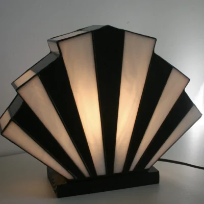 Lampe de table inspirée du style Art Déco des années 1920, en forme de soleil éclatant (appelée 'Sunburst'). Imaginez une structure métallique solide, avec des rayons fins et symétriques qui partent du centre comme les branches d’une étoile, supportant un abat-jour en verre dépoli lisse au toucher. Le verre, légèrement granuleux sous les doigts, adoucit la lumière pour créer une ambiance chaleureuse et tamisée. La base est lourde et stable, avec des courbes géométriques typiques du design rétro. Cette lampe dégage une élégance vintage, comme un objet sorti d’un film en noir et blanc des années 1930.