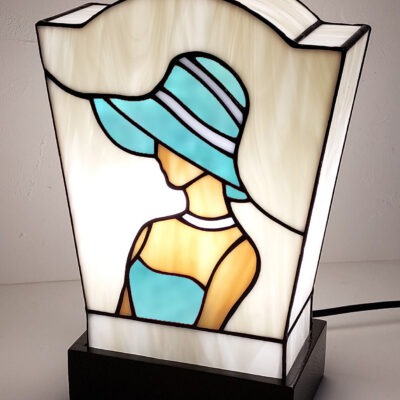 Lampe de table en vitrail de style Art Déco/Tiffany, représentant le profil d'une femme portant un large chapeau à bords. L'abat-jour trapézoïdal est principalement en verre ivoire opalescent. La figure féminine est réalisée avec du verre rouge vif pour la robe et le chapeau (avec des rayures blanches), et du verre ambre pour la peau. Le tout est serti de lignes de plomb noires. La lampe est posée sur un socle noir, et la lumière intérieure met en valeur le dessin graphique typique de l'Art Déco