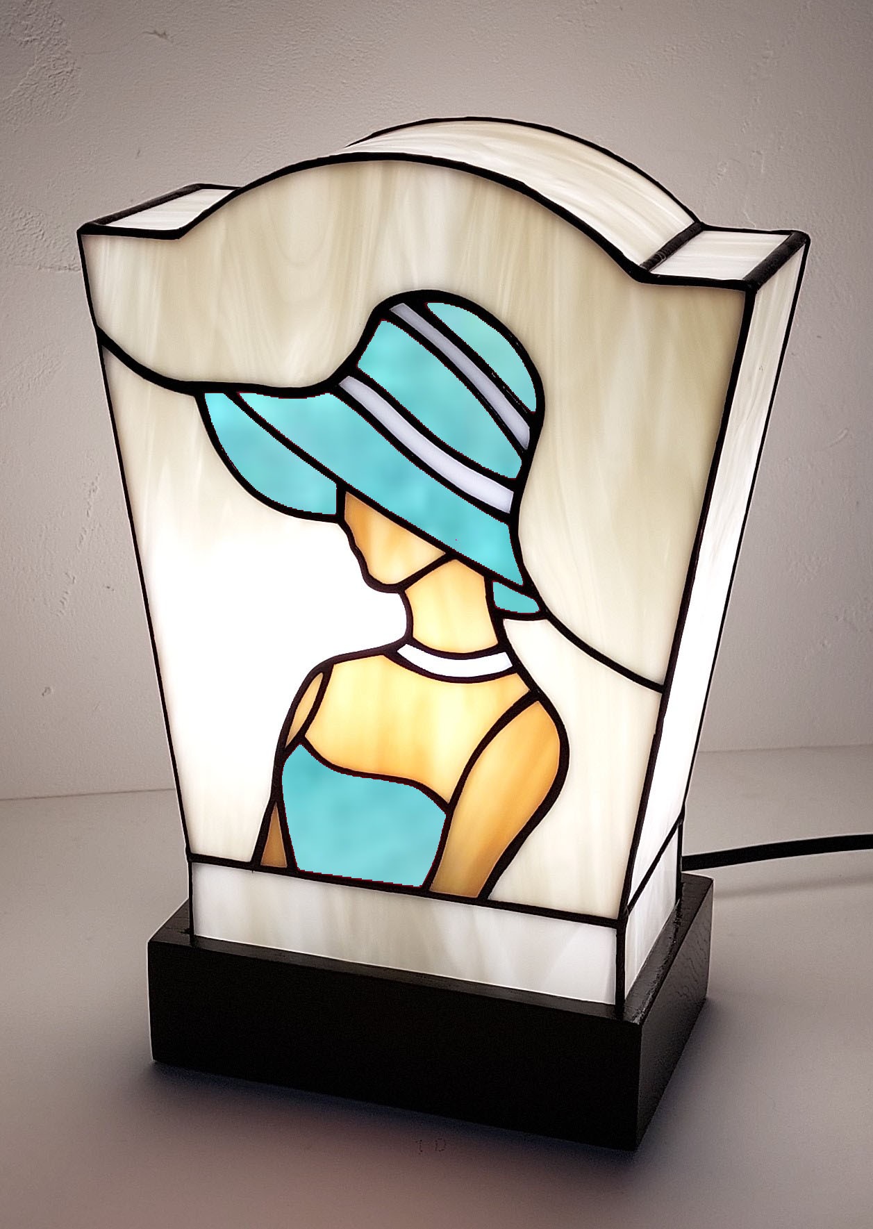 Lampe de table en vitrail de style Art Déco/Tiffany, représentant le profil d'une femme portant un large chapeau à bords. L'abat-jour trapézoïdal est principalement en verre ivoire opalescent. La figure féminine est réalisée avec du verre rouge vif pour la robe et le chapeau (avec des rayures blanches), et du verre ambre pour la peau. Le tout est serti de lignes de plomb noires. La lampe est posée sur un socle noir, et la lumière intérieure met en valeur le dessin graphique typique de l'Art Déco
