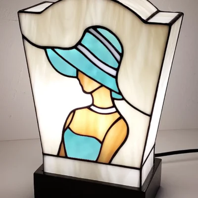Lampe de table en vitrail de style Art Déco/Tiffany, représentant le profil d'une femme portant un large chapeau à bords. L'abat-jour trapézoïdal est principalement en verre ivoire opalescent. La figure féminine est réalisée avec du verre rouge vif pour la robe et le chapeau (avec des rayures blanches), et du verre ambre pour la peau. Le tout est serti de lignes de plomb noires. La lampe est posée sur un socle noir, et la lumière intérieure met en valeur le dessin graphique typique de l'Art Déco